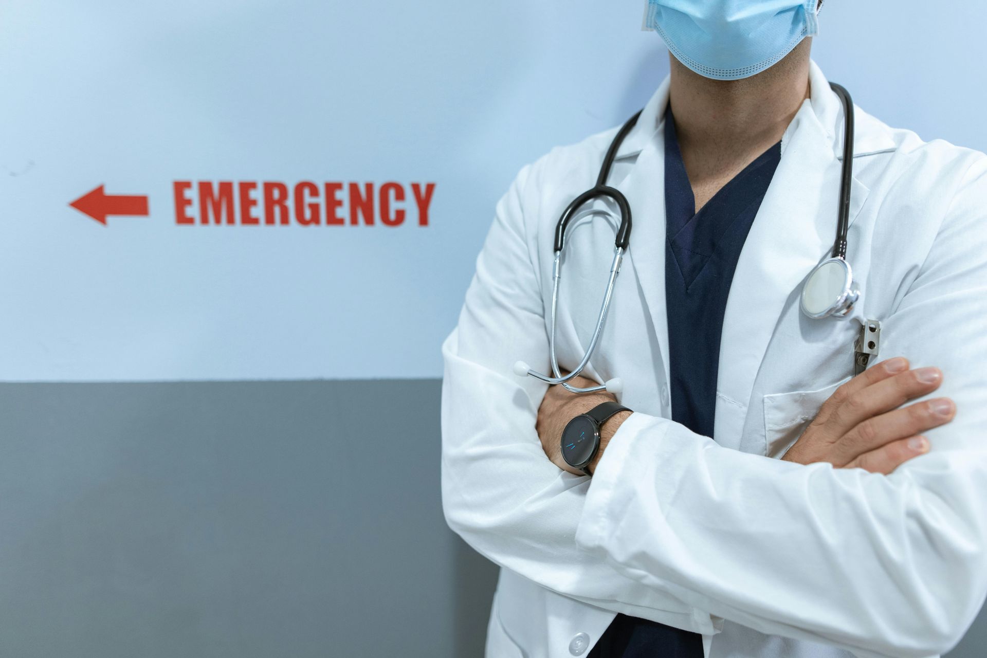 Doctor in white coat and mask, arms crossed, in front of an emergency room sign.
