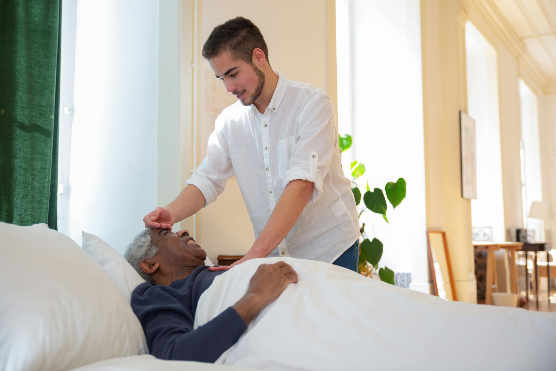Young man tending to an older person in bed, touching their forehead in a sunlit room.