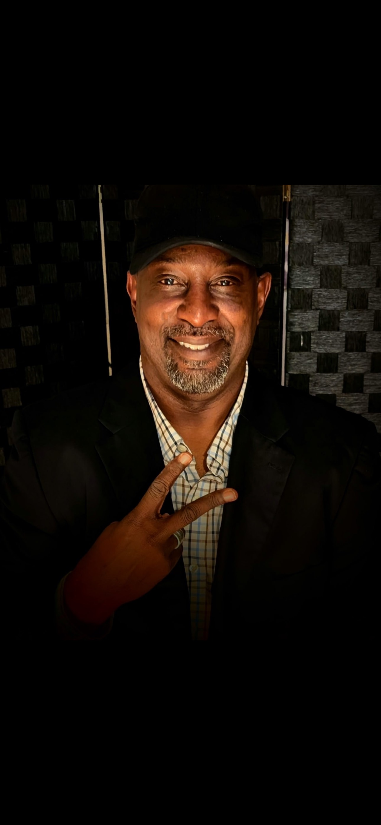 Man in a black hat and blazer smiles and flashes a peace sign. He is in front of a dark, checkered background.