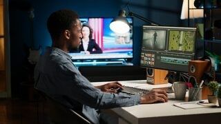 A Black man edits video on a computer in a dark room, with a TV showing news in the background.
