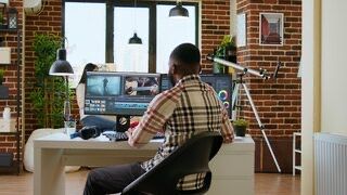 Man in plaid shirt editing video on dual monitors in a brick-walled room, desk with equipment, telescope in the corner.
