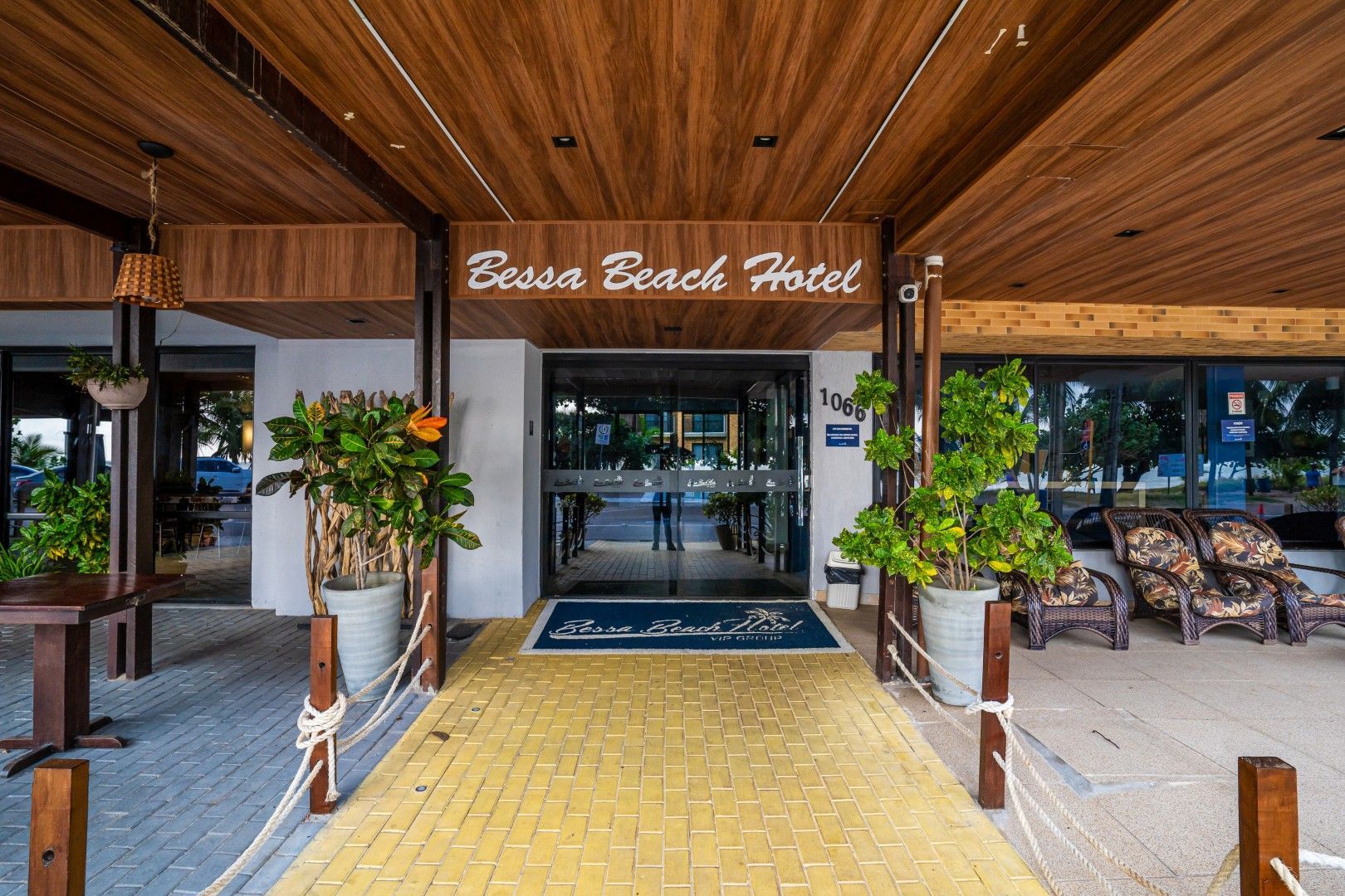 Entrada do Hotel Esma Beach com cobertura de madeira, passarela amarela e vasos de plantas.