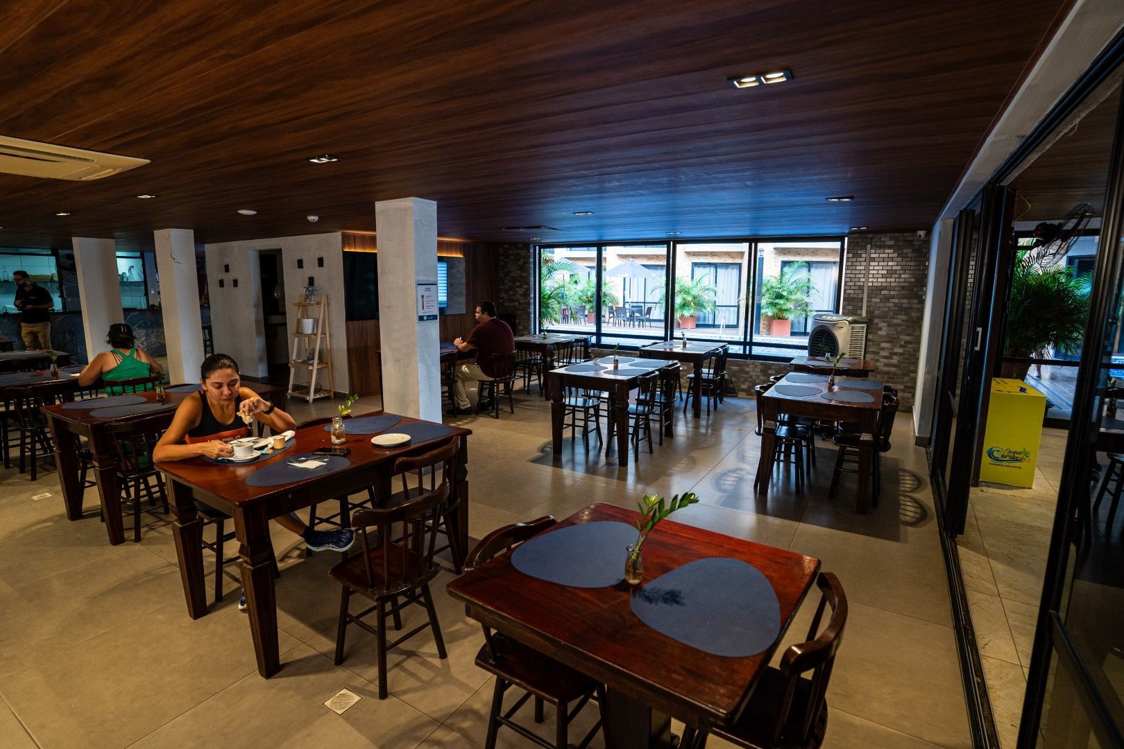 Sala de jantar espaçosa do restaurante com mesas de madeira, grandes janelas e alguns lugares para clientes sentarem-se.