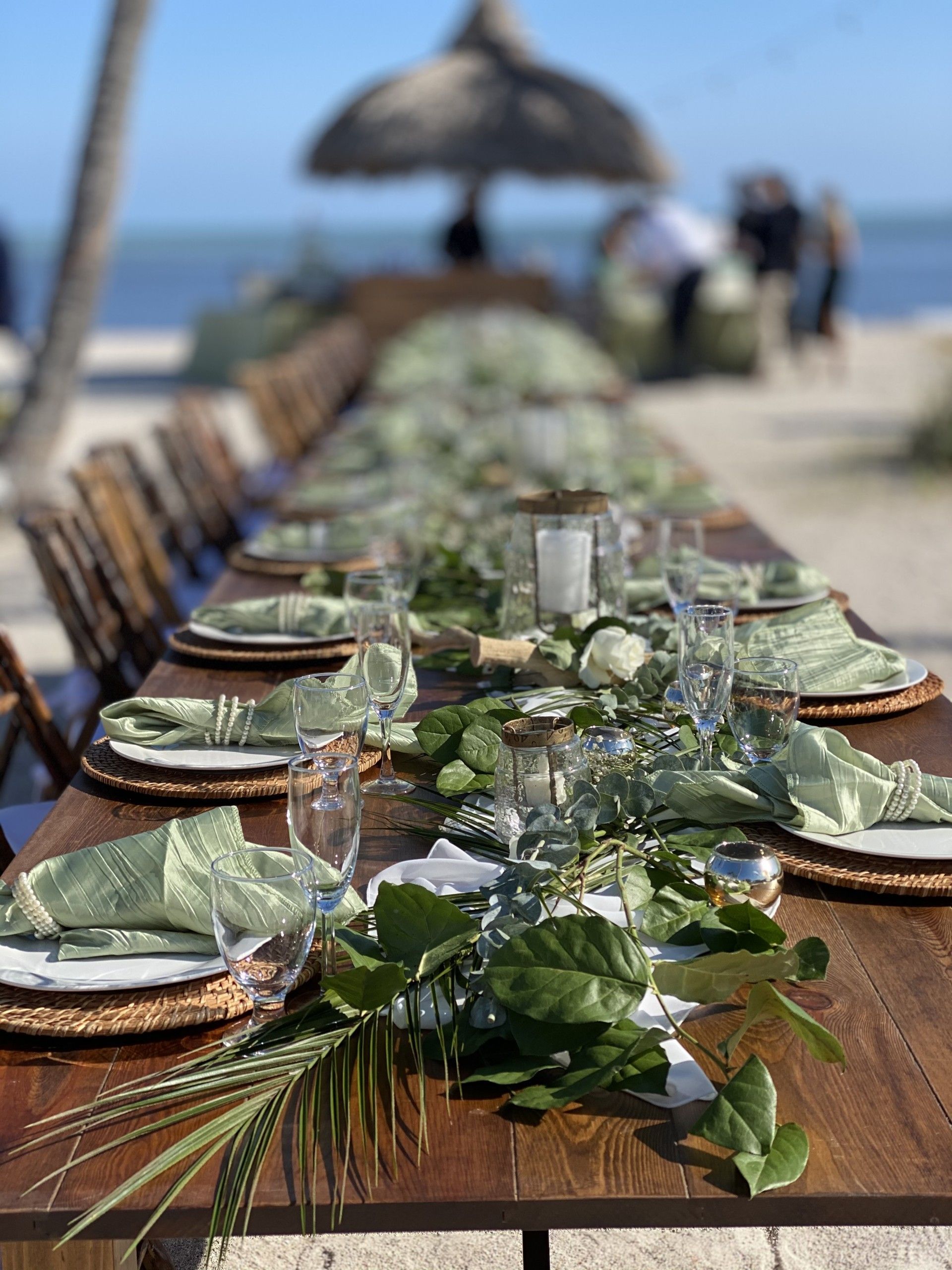 Long wooden table set for dining on a beach, decorated with greenery and place settings.