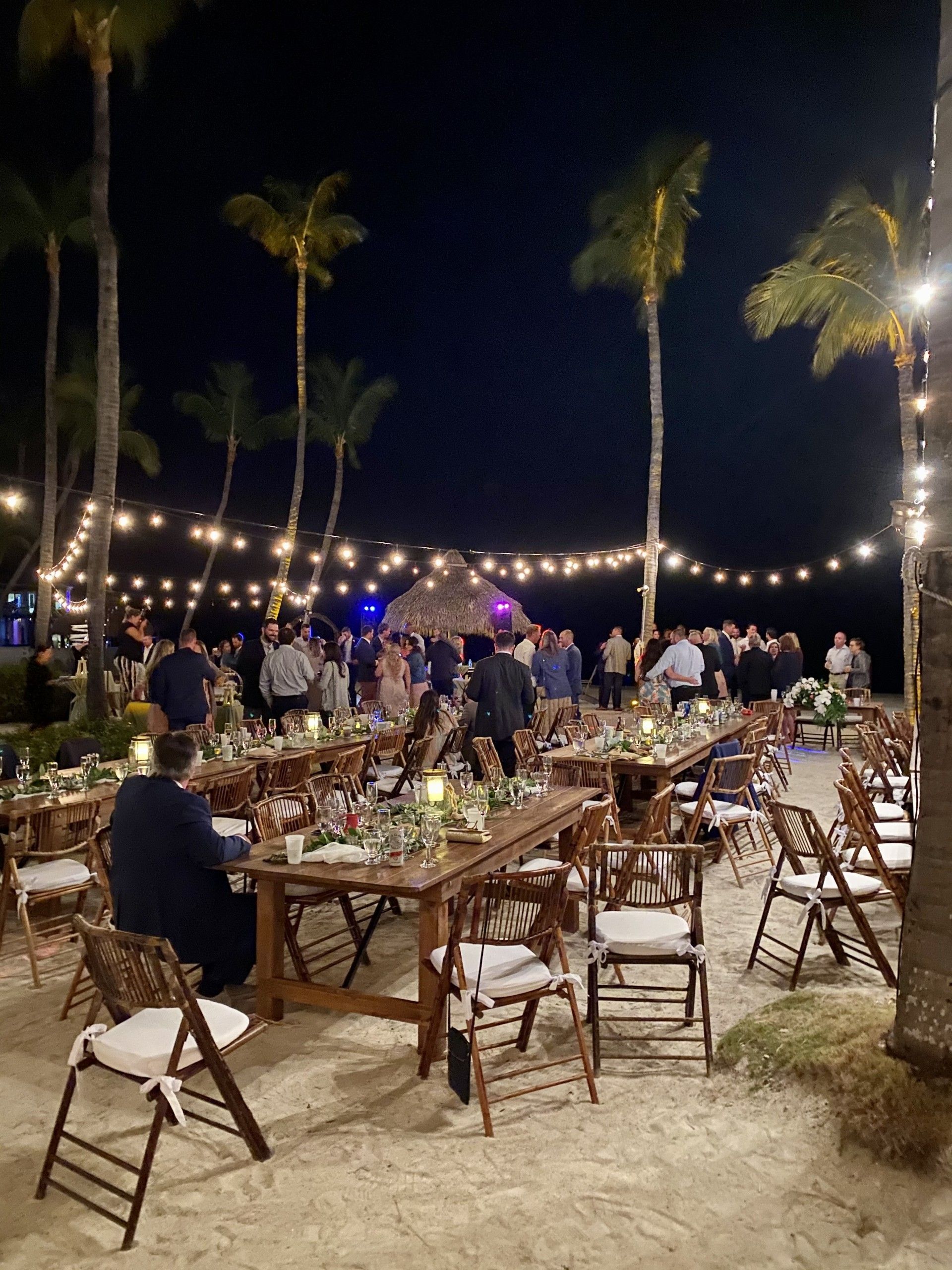 Outdoor reception at night, tables set for dinner on beach, string lights, palm trees, guests socializing.