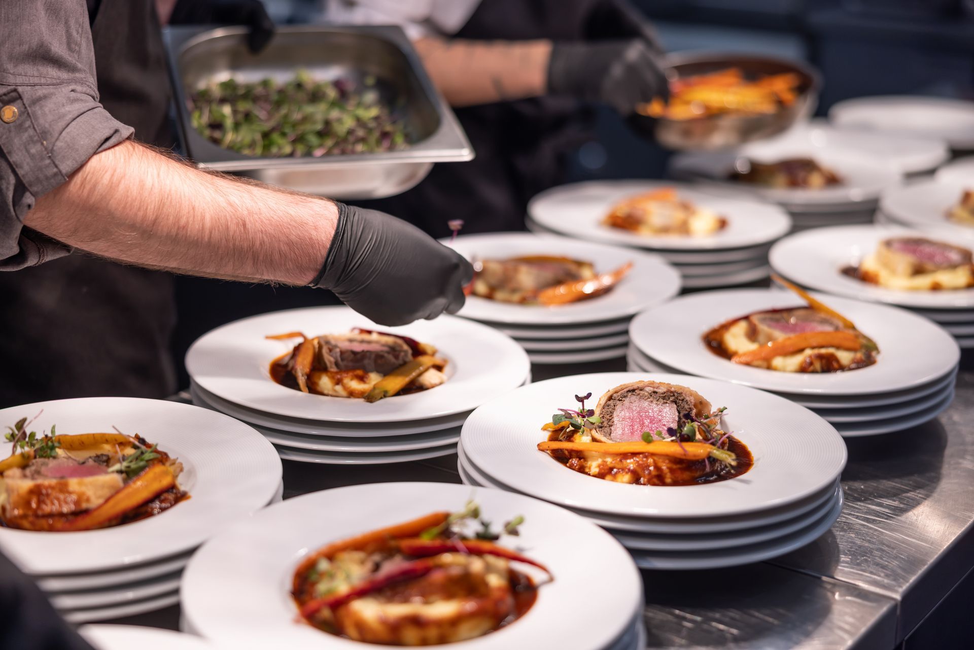 Chefs plating gourmet dishes with meat, carrots, and greens on white plates in a restaurant kitchen.