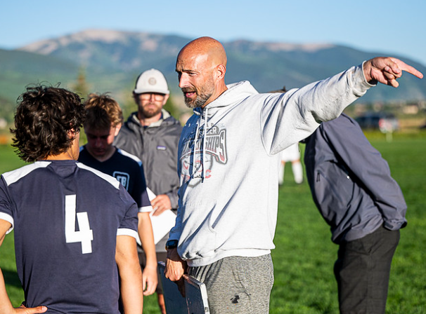 a man wearing a sweatshirt with the number 4 on it is talking to a group of boys on a field .