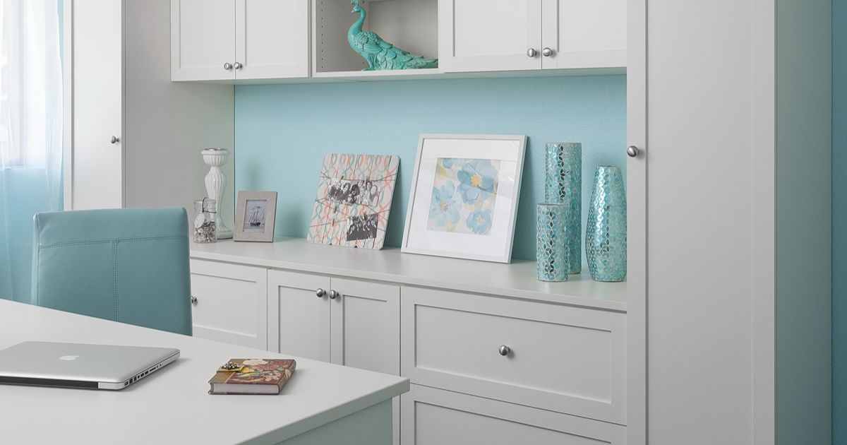A home office with white built-in cabinets, light blue accents, a teal office chair, and a laptop on a white desk.