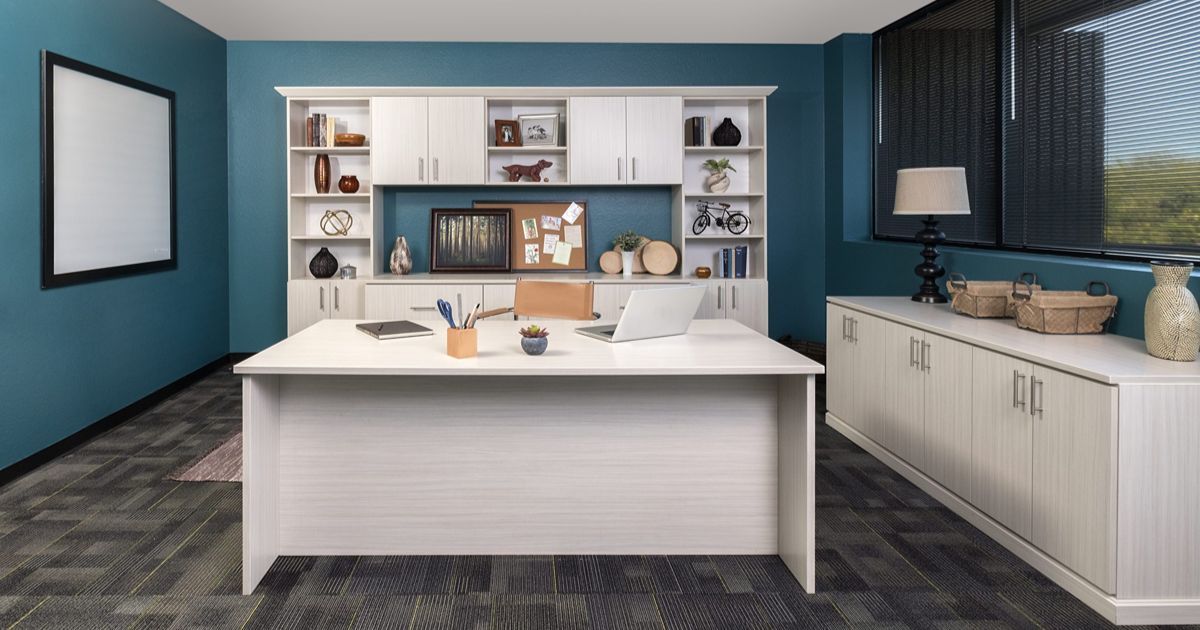 A modern office with a white desk, matching storage cabinetry, teal walls, and dark patterned flooring.