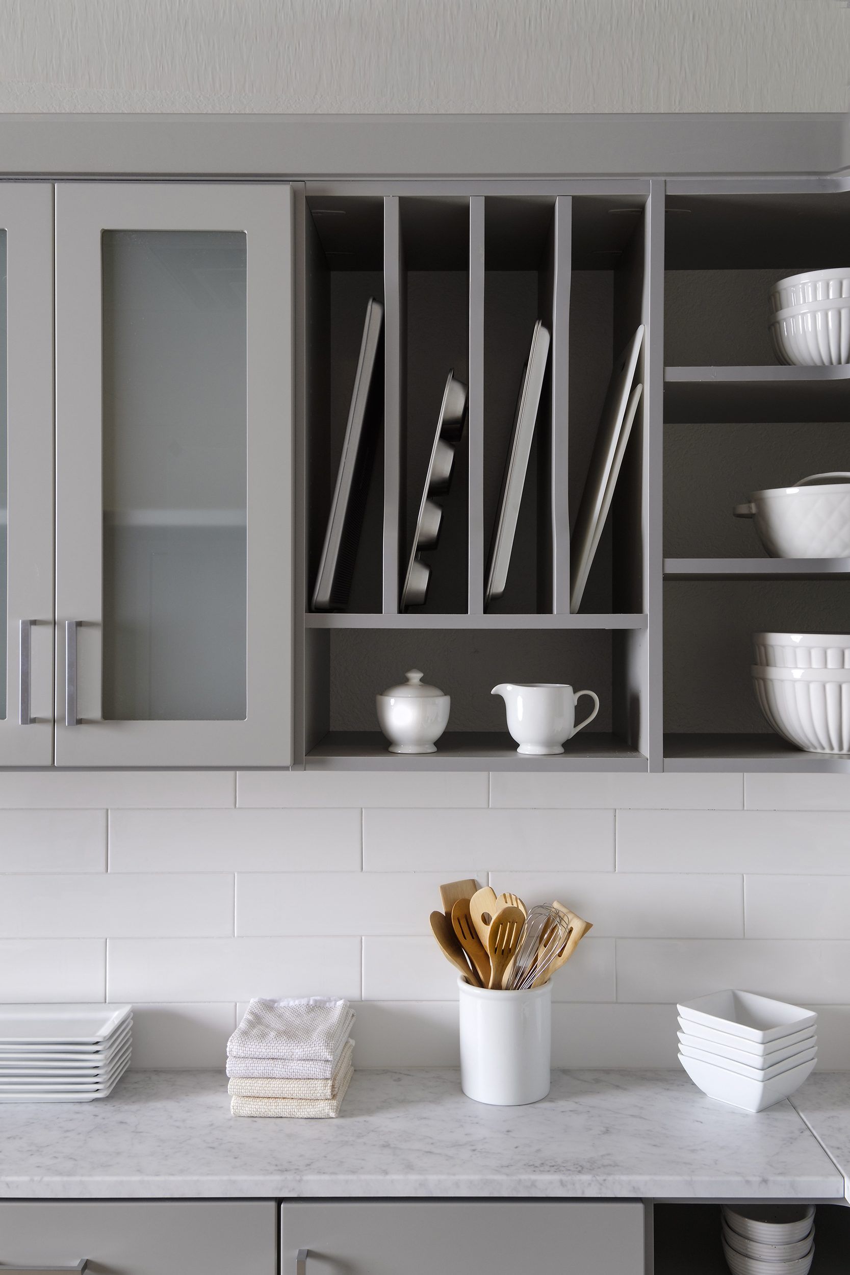 Modern kitchen cabinets with vertical plate storage, open shelves, and a white countertop with kitchen tools and dishes.