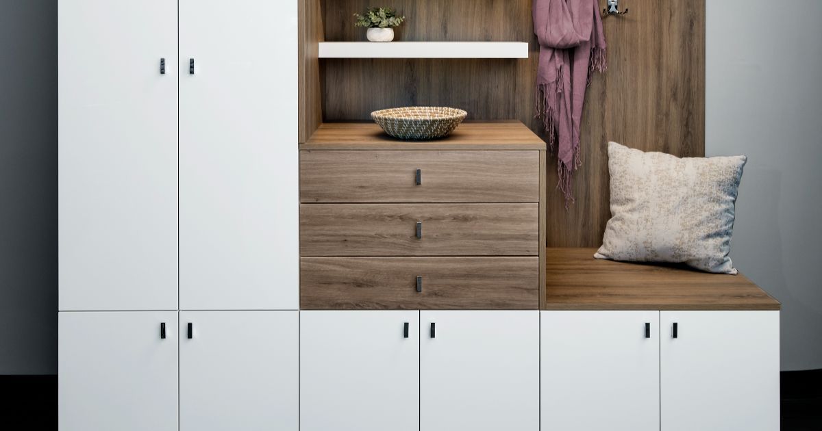 A mudroom storage unit featuring white cabinets, wood-tone drawers, a built-in bench with a throw pillow, and a shelf.