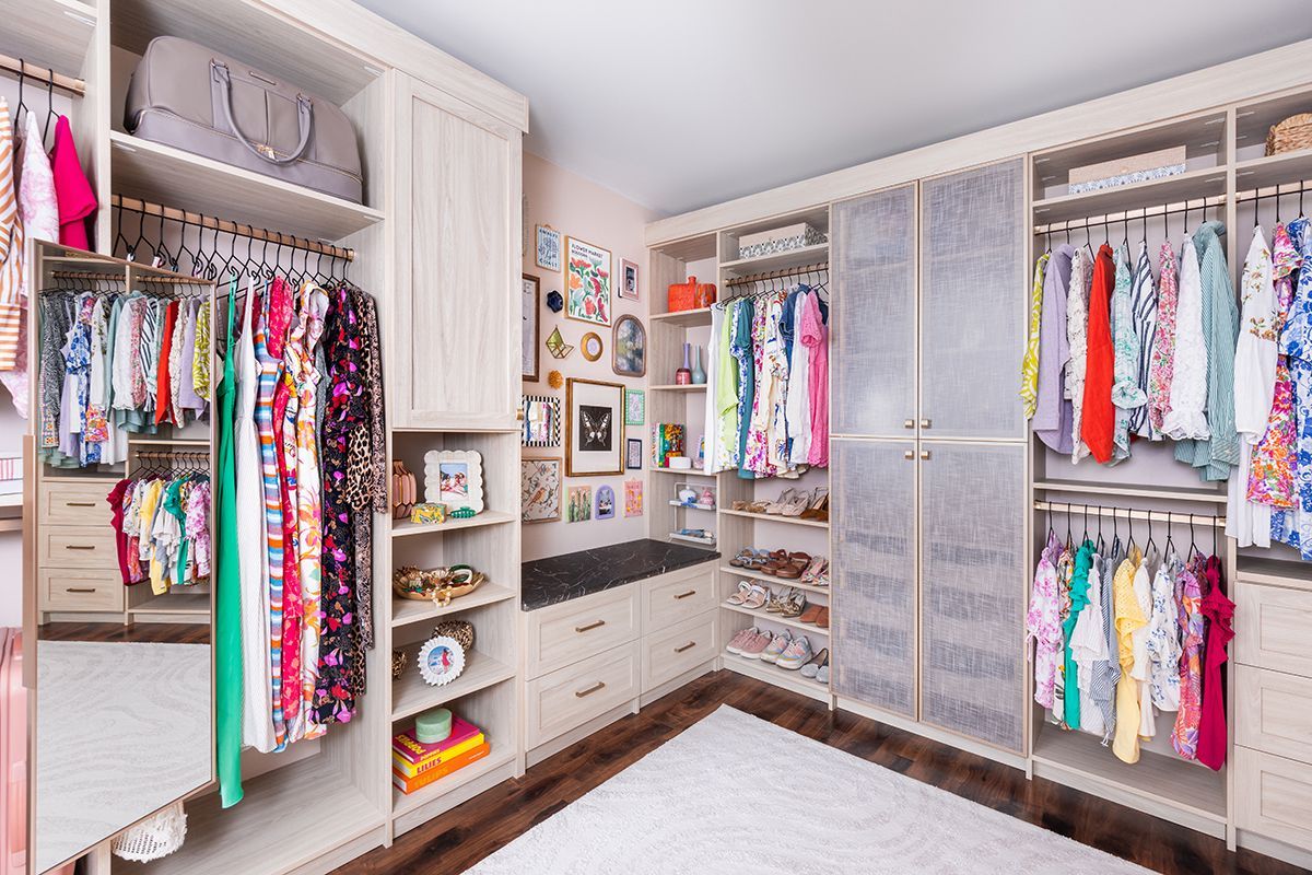 A well-organized walk-in closet featuring light-toned cabinetry, hanging clothes, shelving, and a small gallery wall.