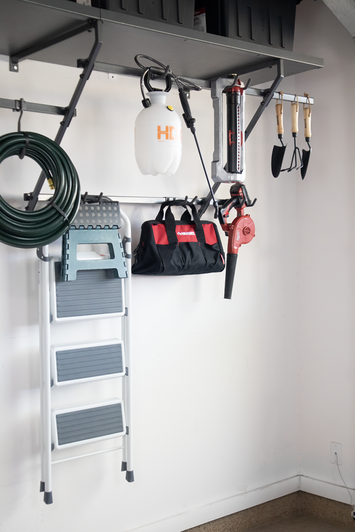 A wall-mounted garage storage system holding a garden hose, sprayer, tool bag, blower, step ladder, and hand tools.