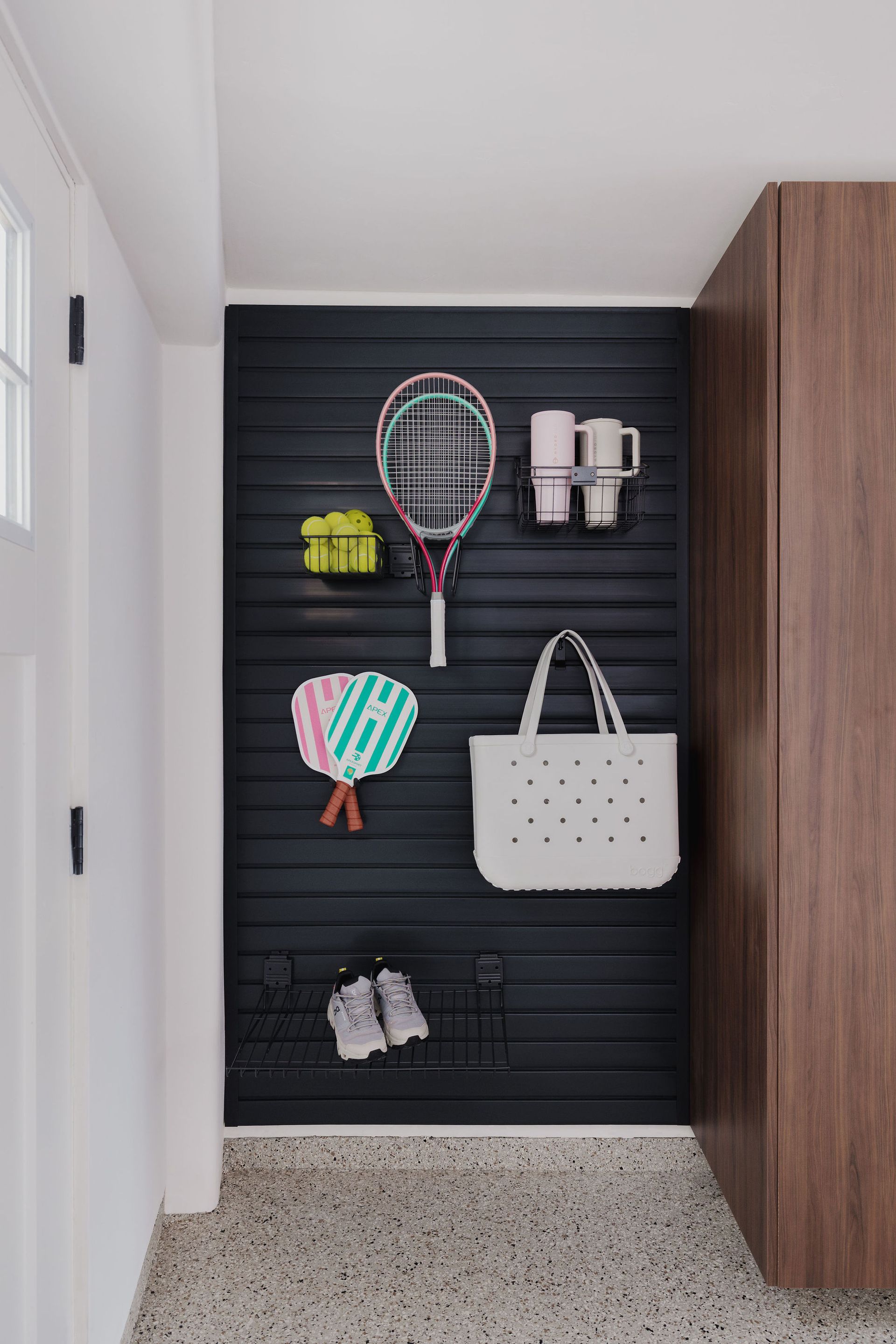 A black slat-wall organizer on an entryway wall holds a tennis racket, balls, paddles, a white tote bag, cups, and shoes.