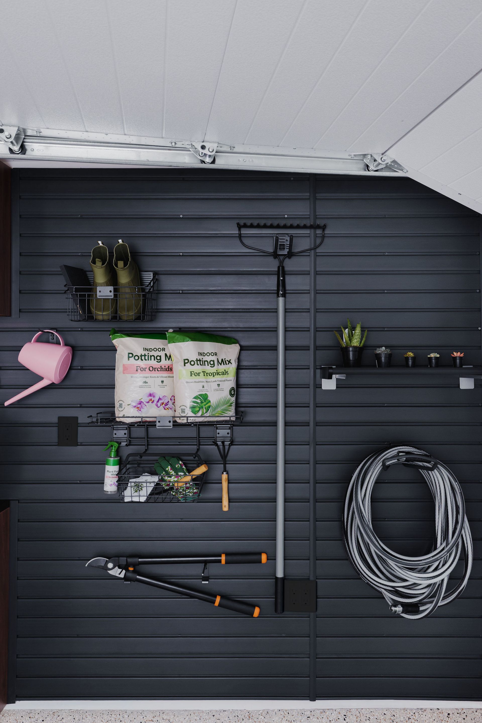Gardening tools and supplies are organized on a dark grey slatted wall, including a hose, pruners, and bags of soil.