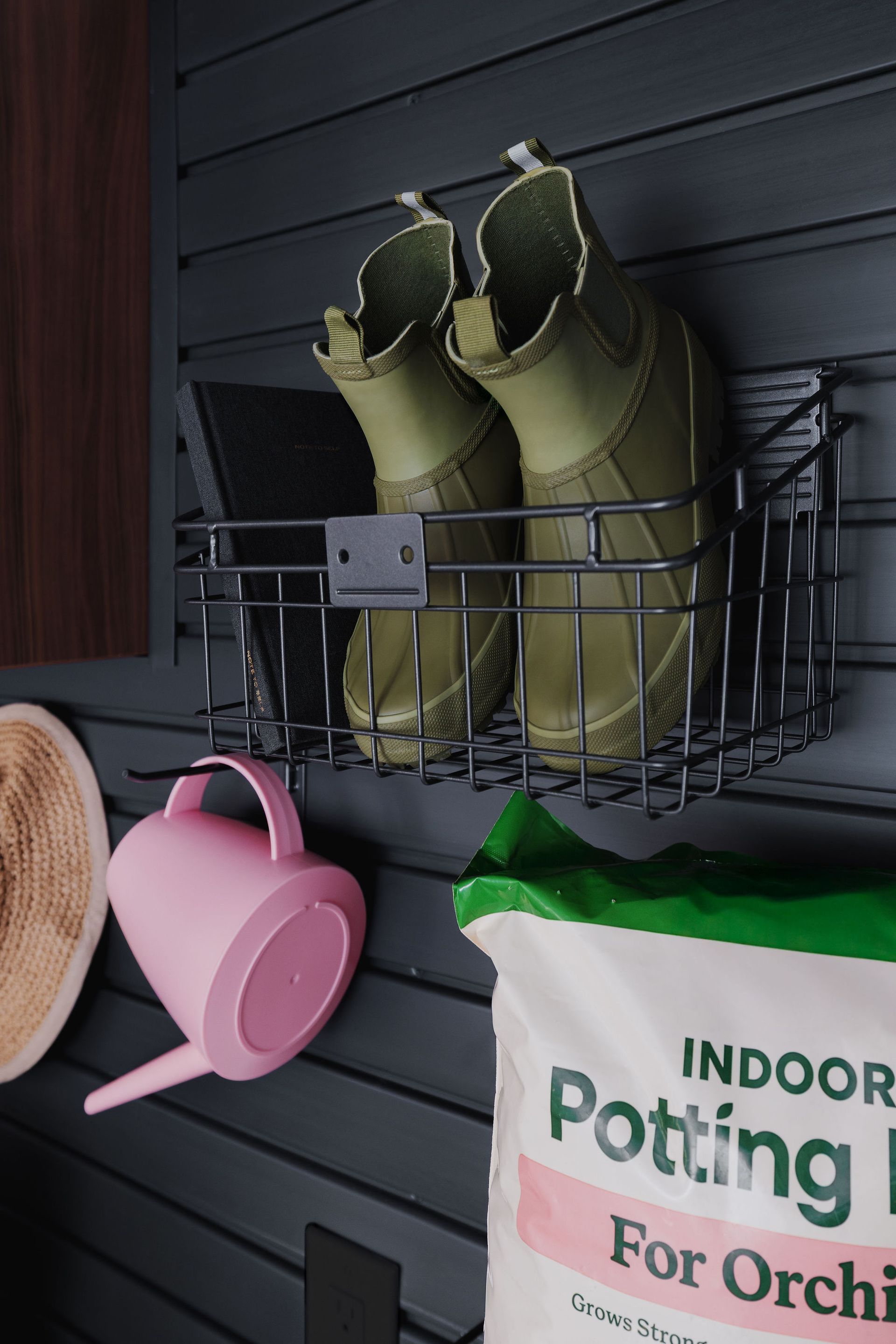 A black slatted wall display holds a wire basket with olive green boots, a pink watering can, and a bag of potting mix.