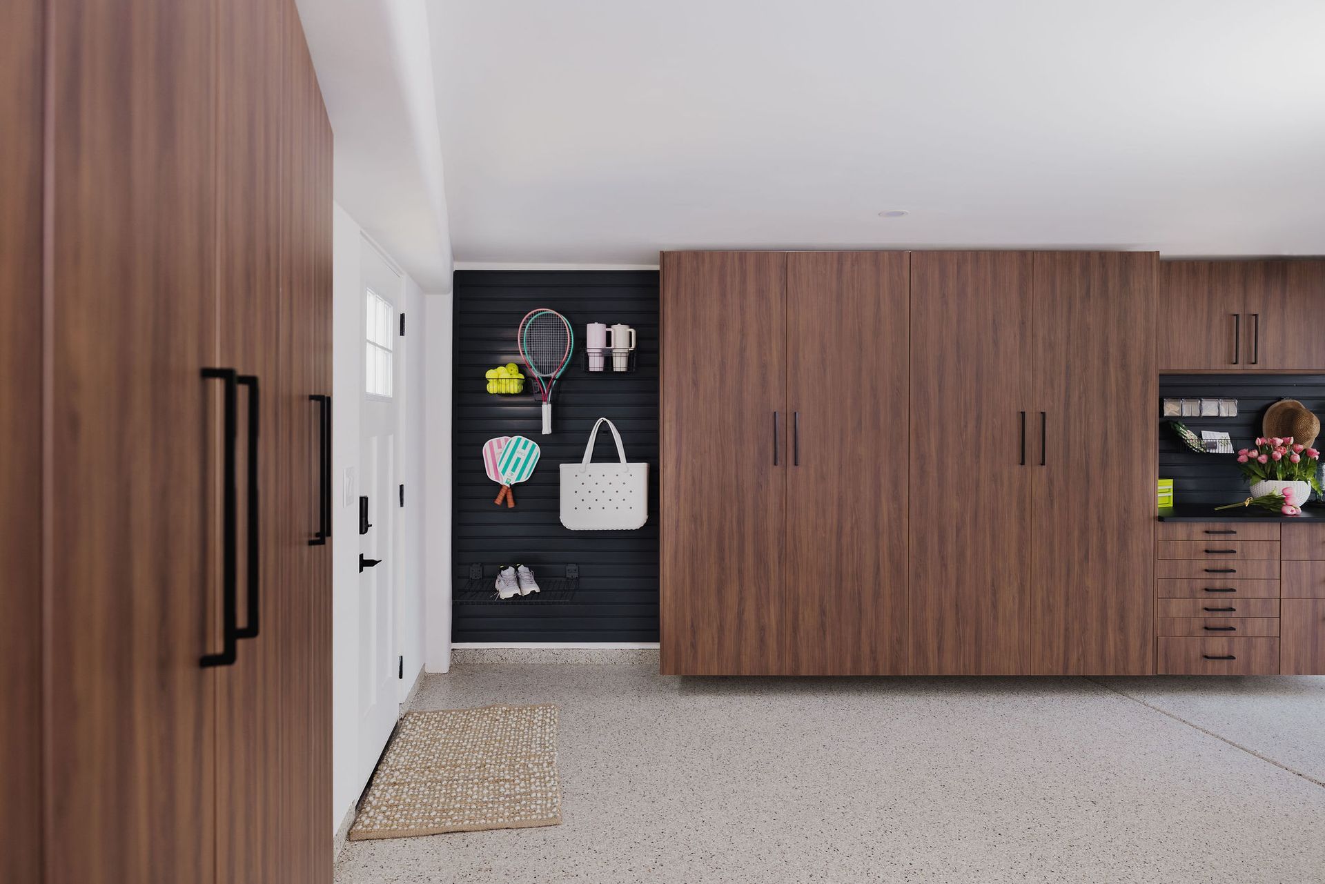 Modern garage interior with wood-grain cabinets, a dark slat wall organizer for sports gear, and speckled floor tiles.