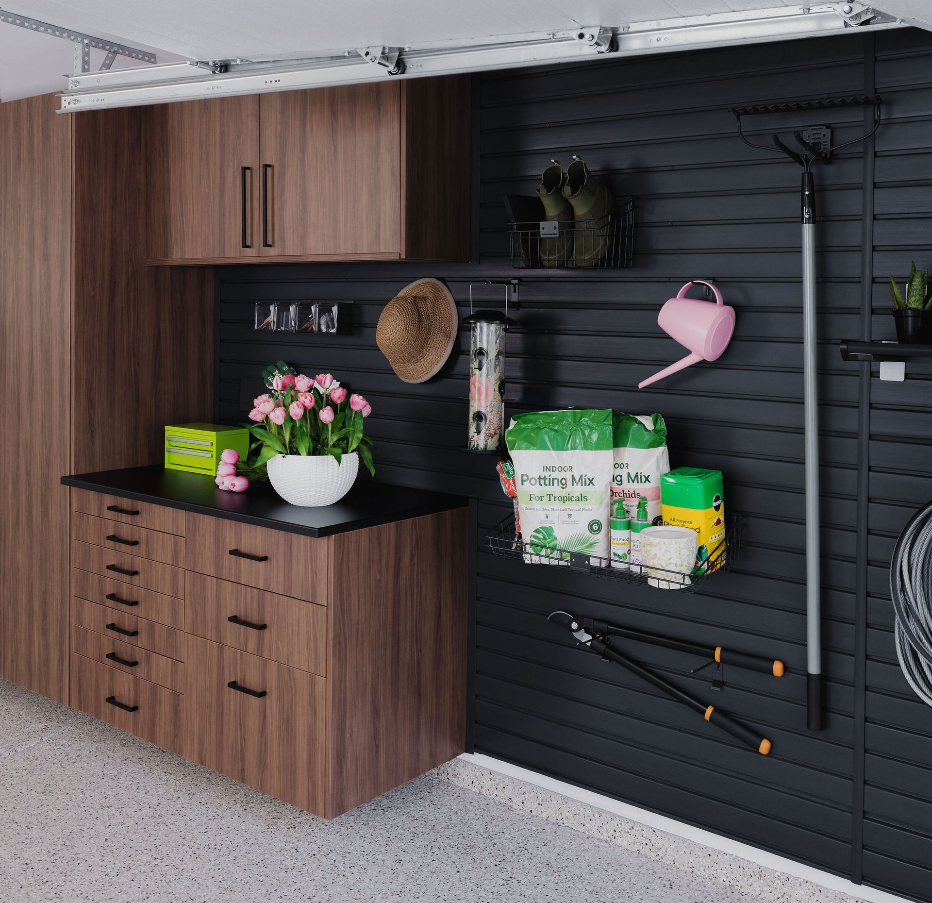 A modern garage workspace featuring dark wood cabinets, black slat wall storage, gardening tools, and potted flowers.