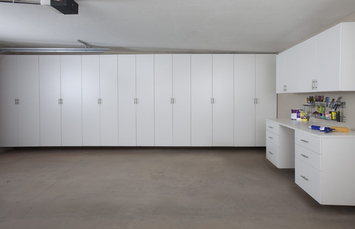 White garage storage cabinets line a wall next to a workbench with drawers and a wall-mounted tool rack.