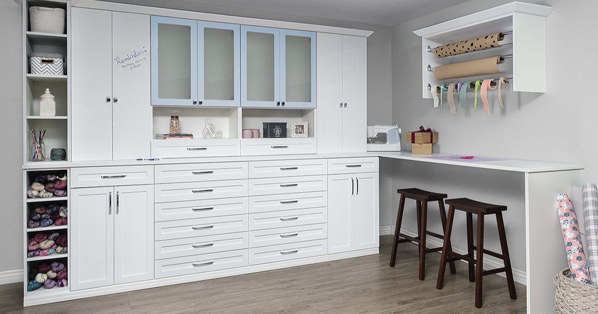 A well-organized craft room featuring white cabinetry, frosted glass cupboards, built-in drawers, and a wrapping paper rack.