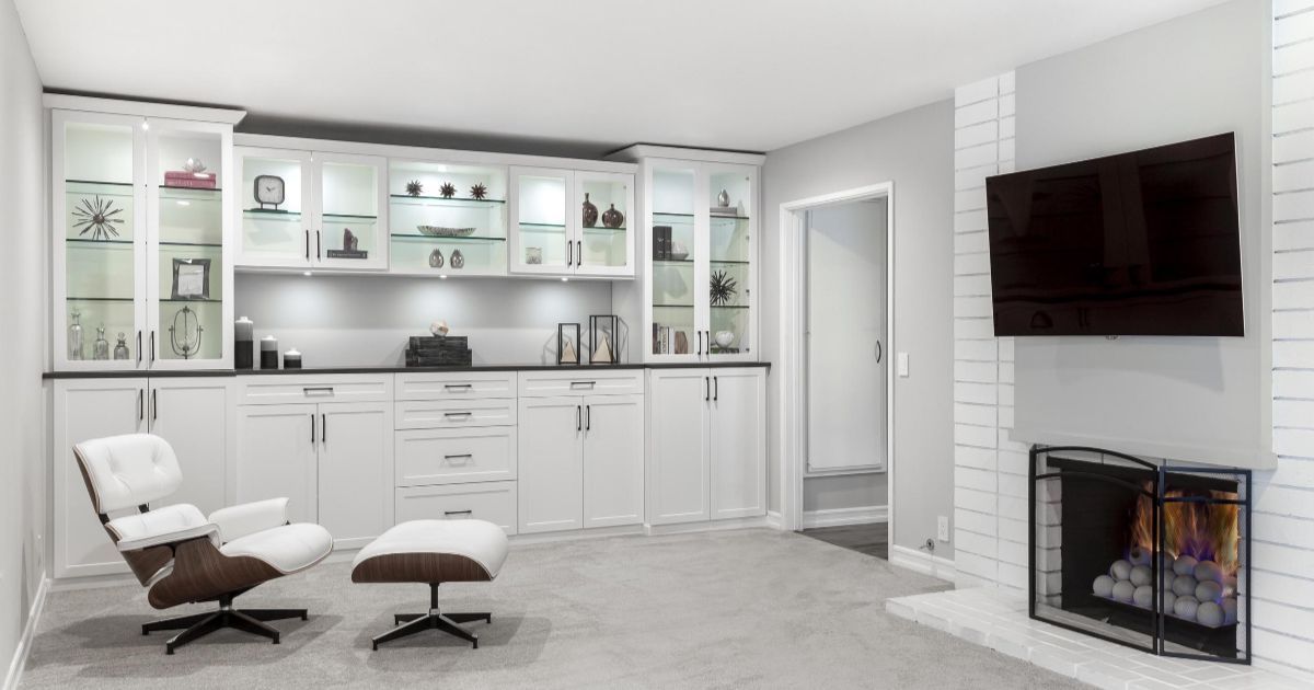 A white built-in cabinet and lounge chair are positioned beside a fireplace with a mounted television in a bright room.