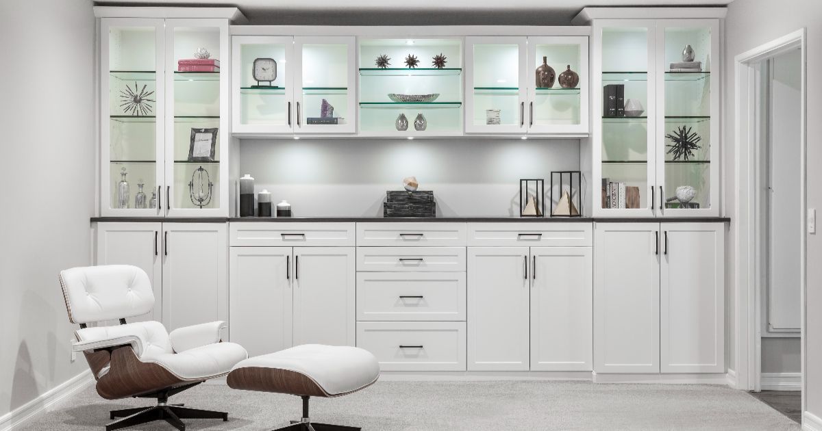 A modern white built-in cabinetry wall with glass-front displays, interior lighting, and a lounge chair with an ottoman.