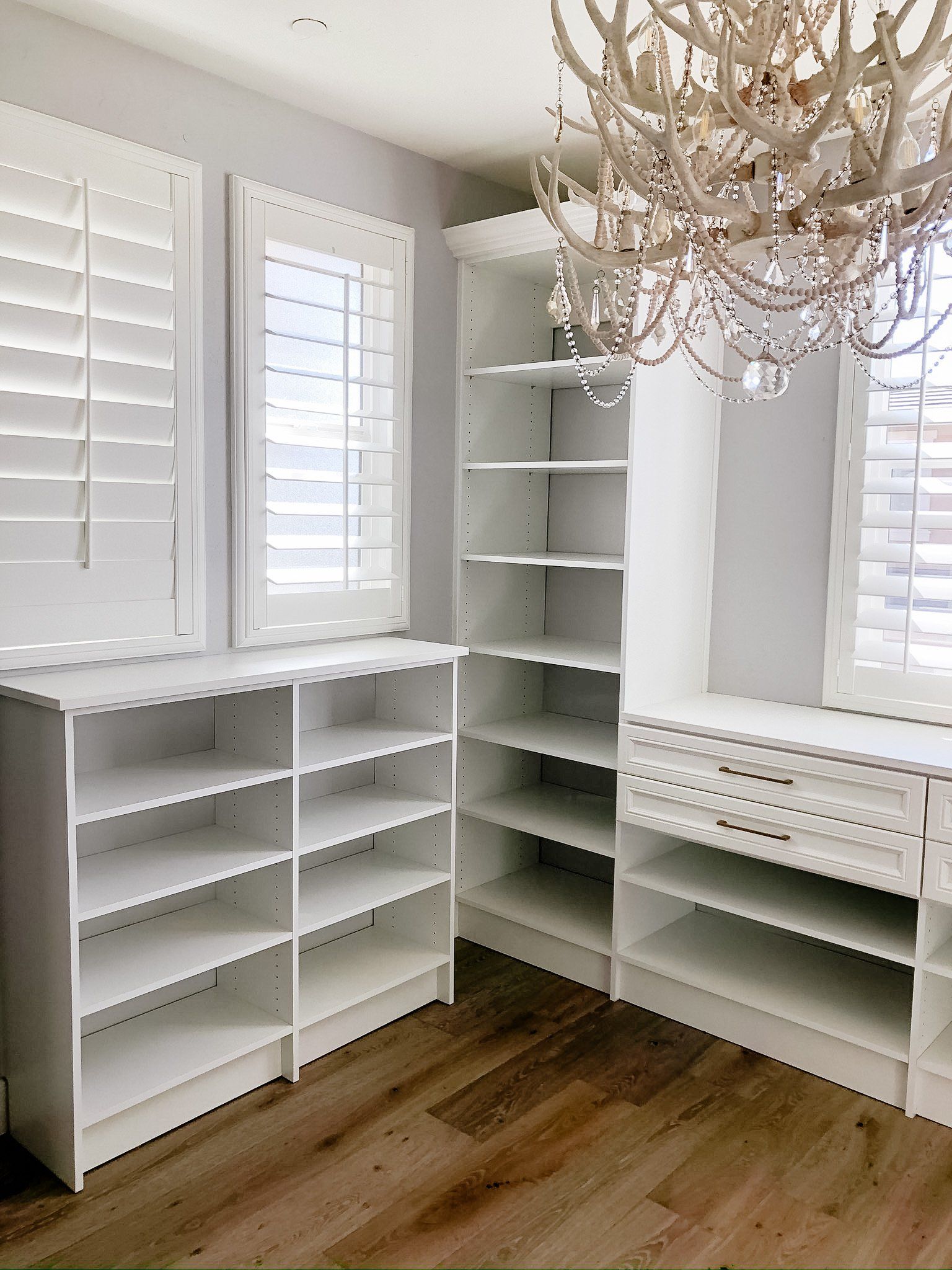 A walk in closet with white shelves and shutters and a chandelier.