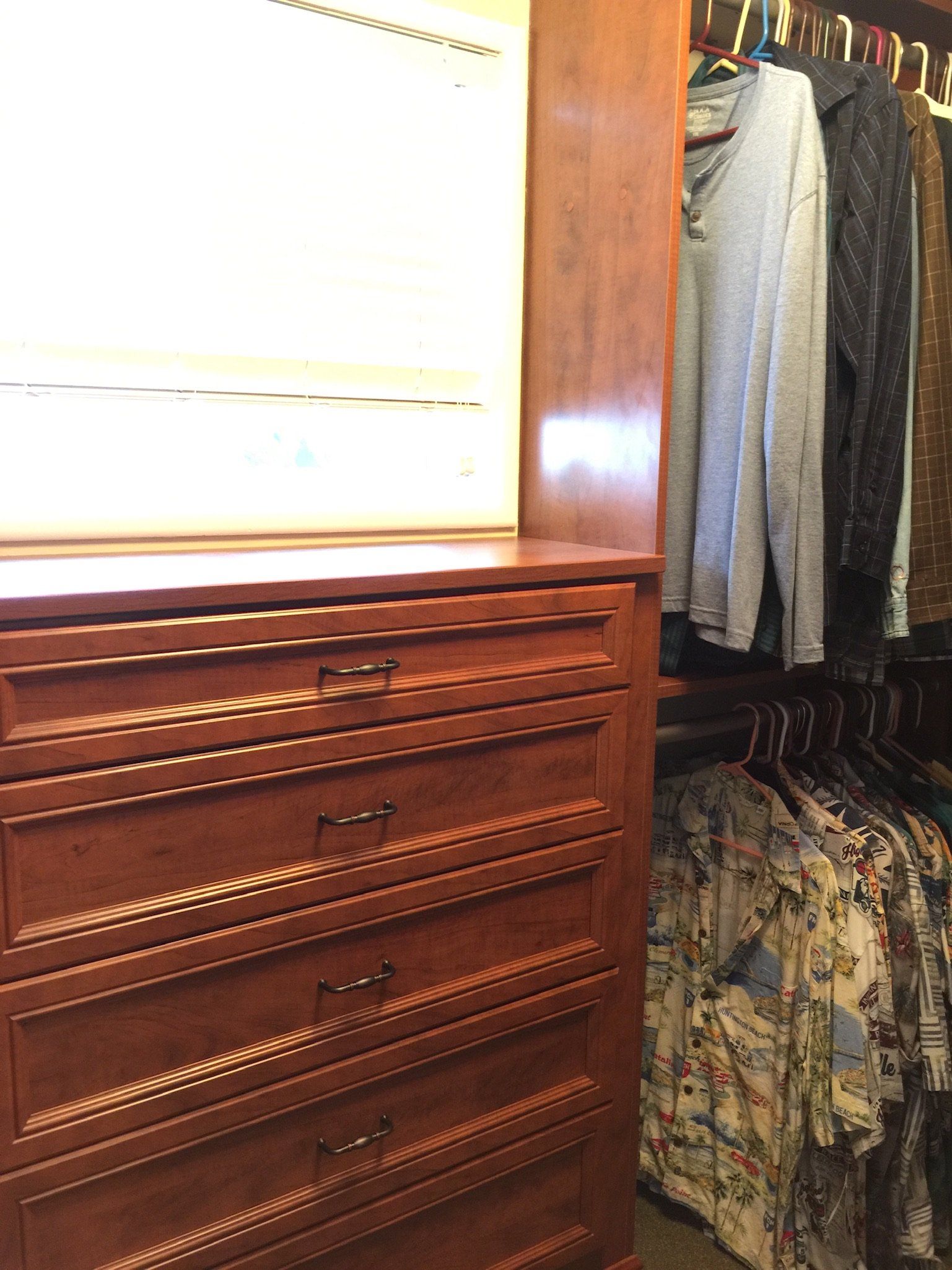 A wooden dresser in a walk in closet filled with clothes.
