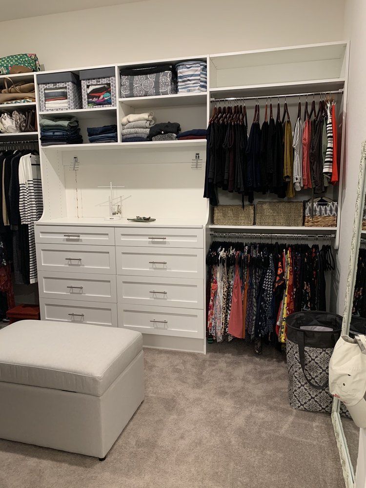 A walk in closet with lots of clothes and a white ottoman.