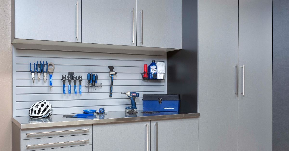 A modern grey garage storage system featuring cabinets, a stainless steel countertop, a slat-wall organizer, and tools.