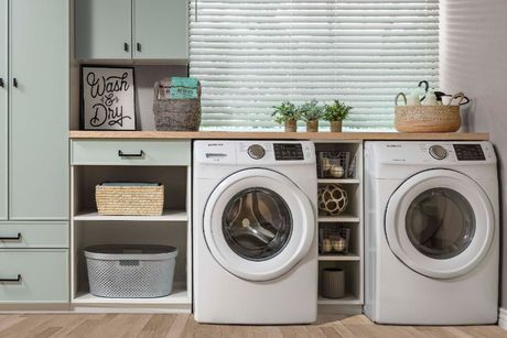 A laundry room with white washer and dryer, sage green cabinets, light wood countertop, and organized storage baskets.