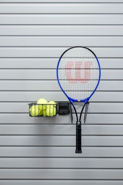 A tennis racket is hanging on a wall next to a basket of tennis balls.