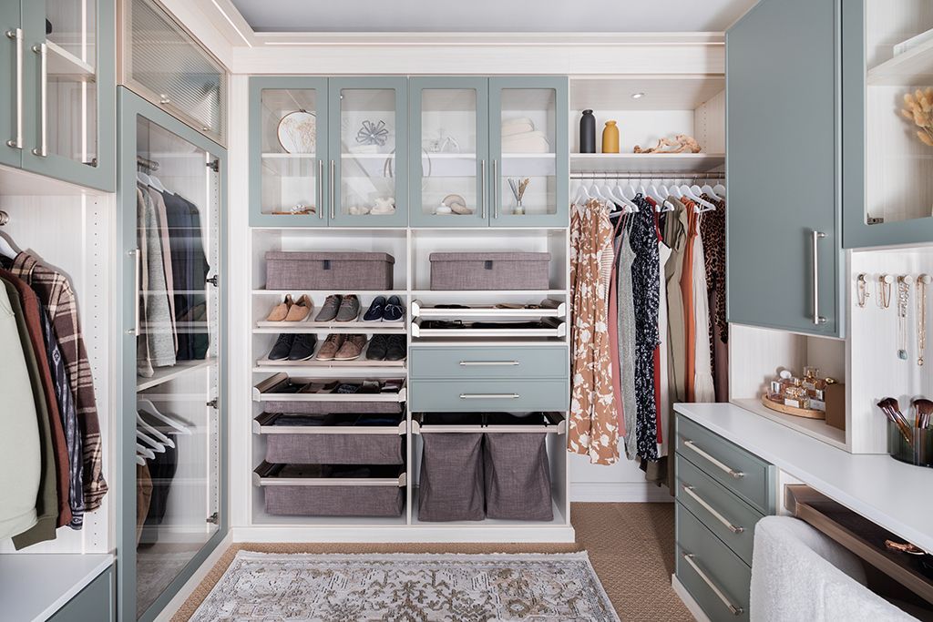 A walk-in closet with sage green cabinets, hanging clothes, drawers, shelves, storage baskets, and a central rug.