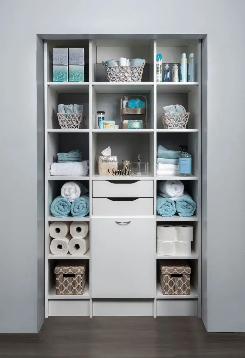 A white, built-in bathroom storage closet with shelves holding folded blue and white towels, toilet paper, and baskets.