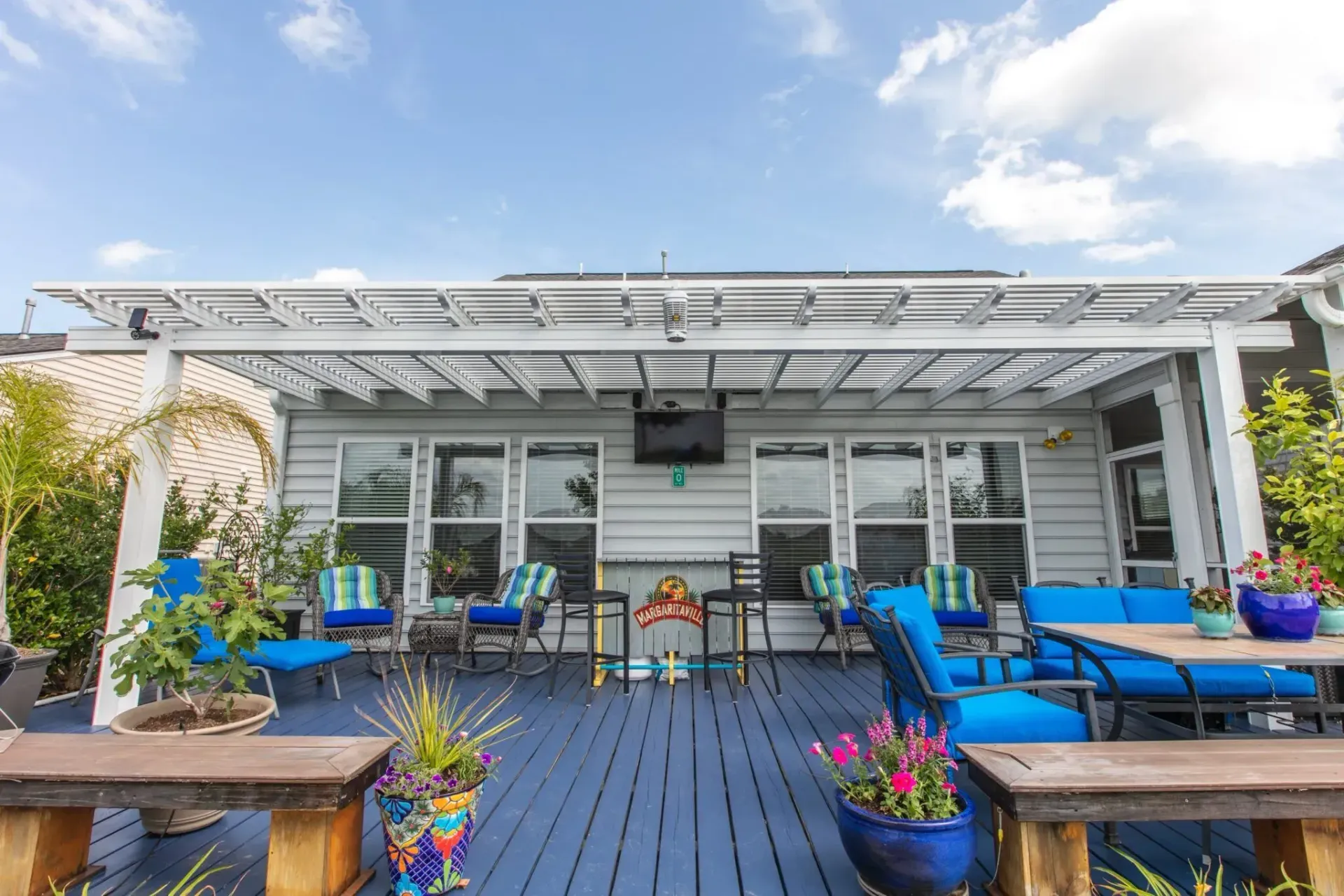 Outdoor patio with blue chairs, tables, and potted flowers under a white pergola by a building facade