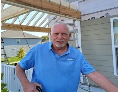 Jim in a blue shirt is standing in front of a house holding a tape measure .