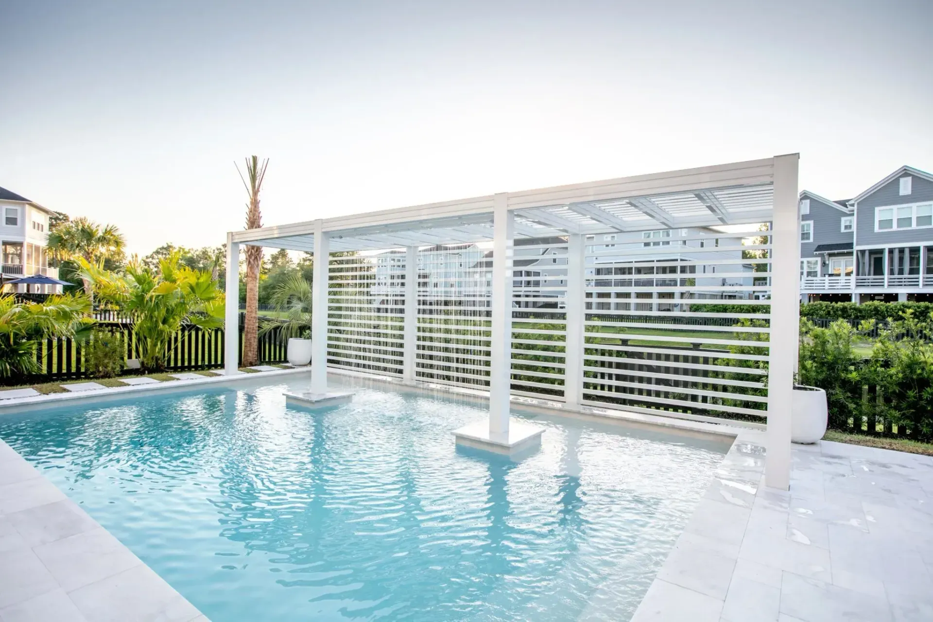 Outdoor swimming pool with a white pergola and suburban homes in the background