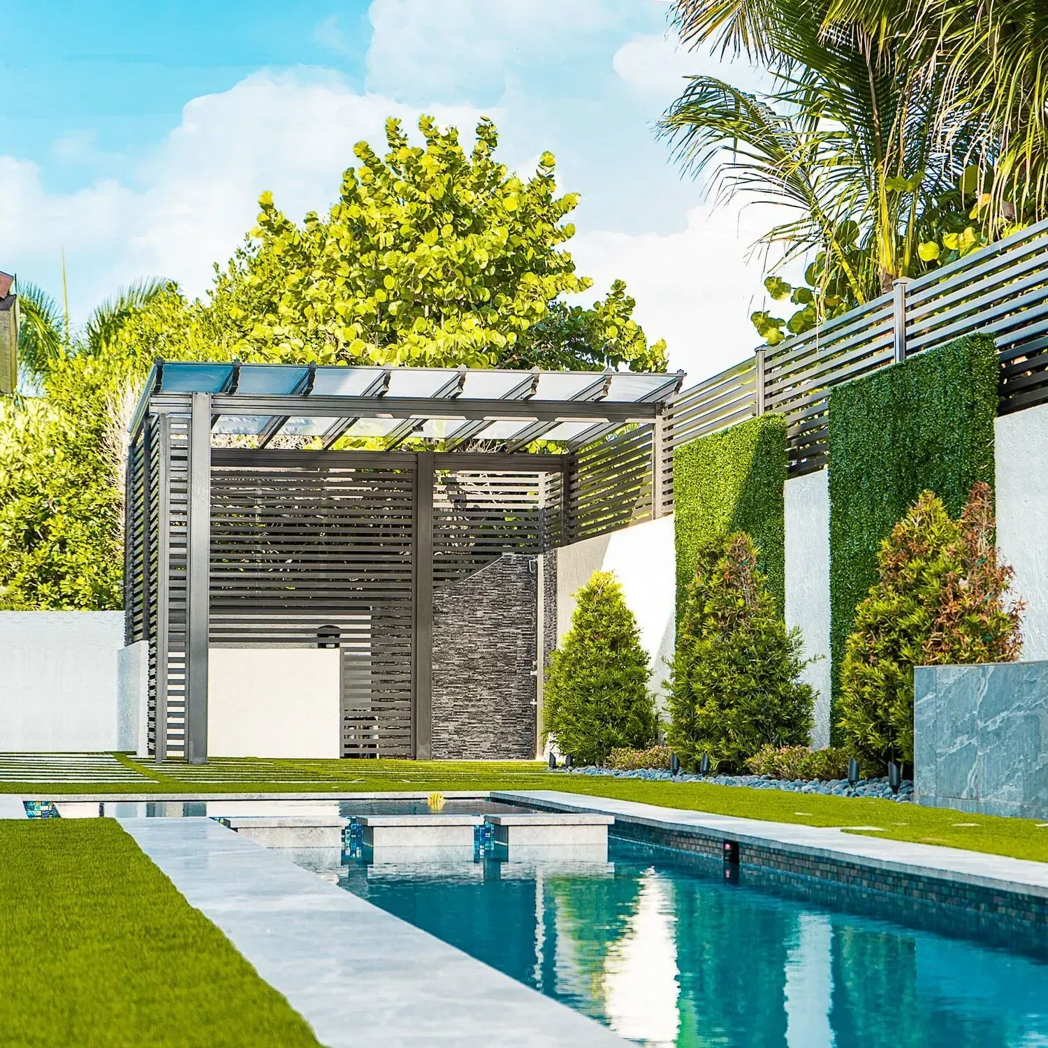 Modern backyard pool with pergola, tiled water feature, and manicured greenery under a bright sky