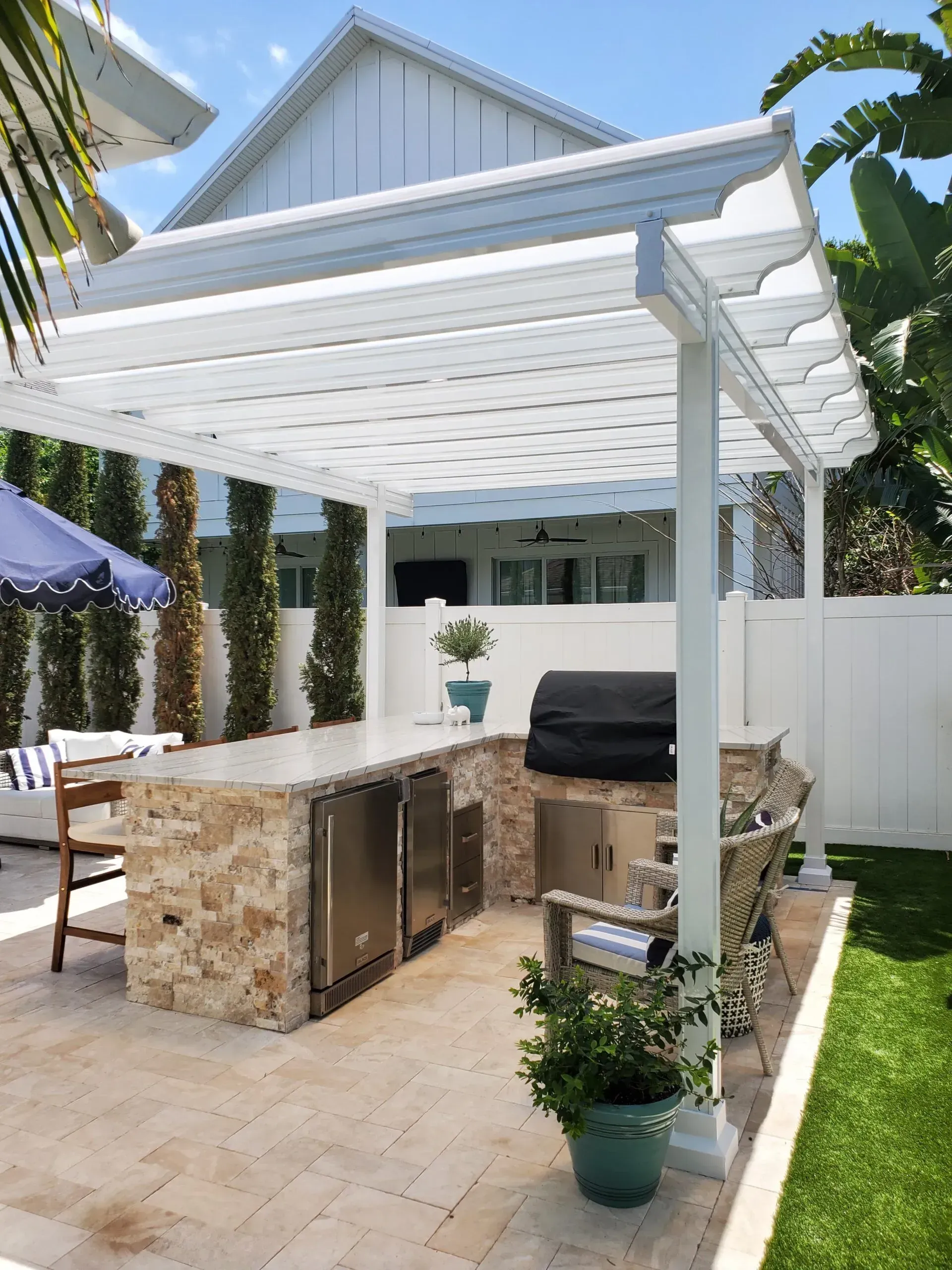 Patio with stone outdoor kitchen under a white pergola beside a house and green lawn