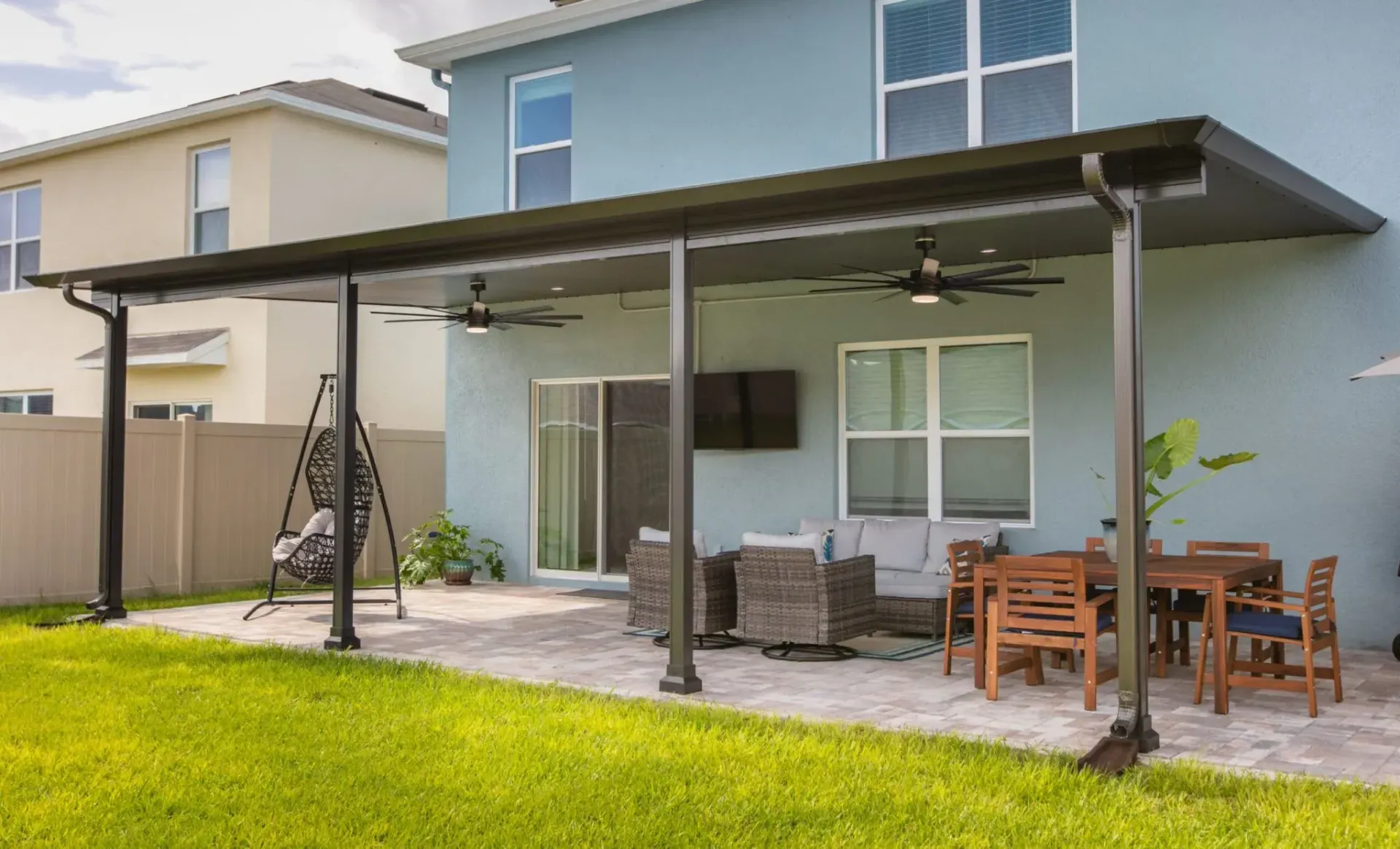 Backyard patio with covered seating, outdoor dining table, and green lawn beside a two-story house