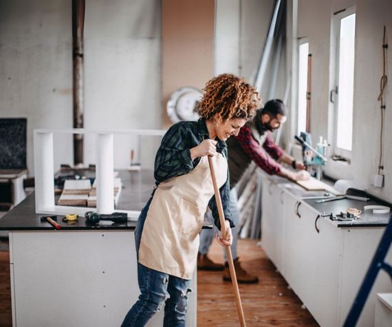 A woman is sweeping the floor in a kitchen while a man works on a counter.