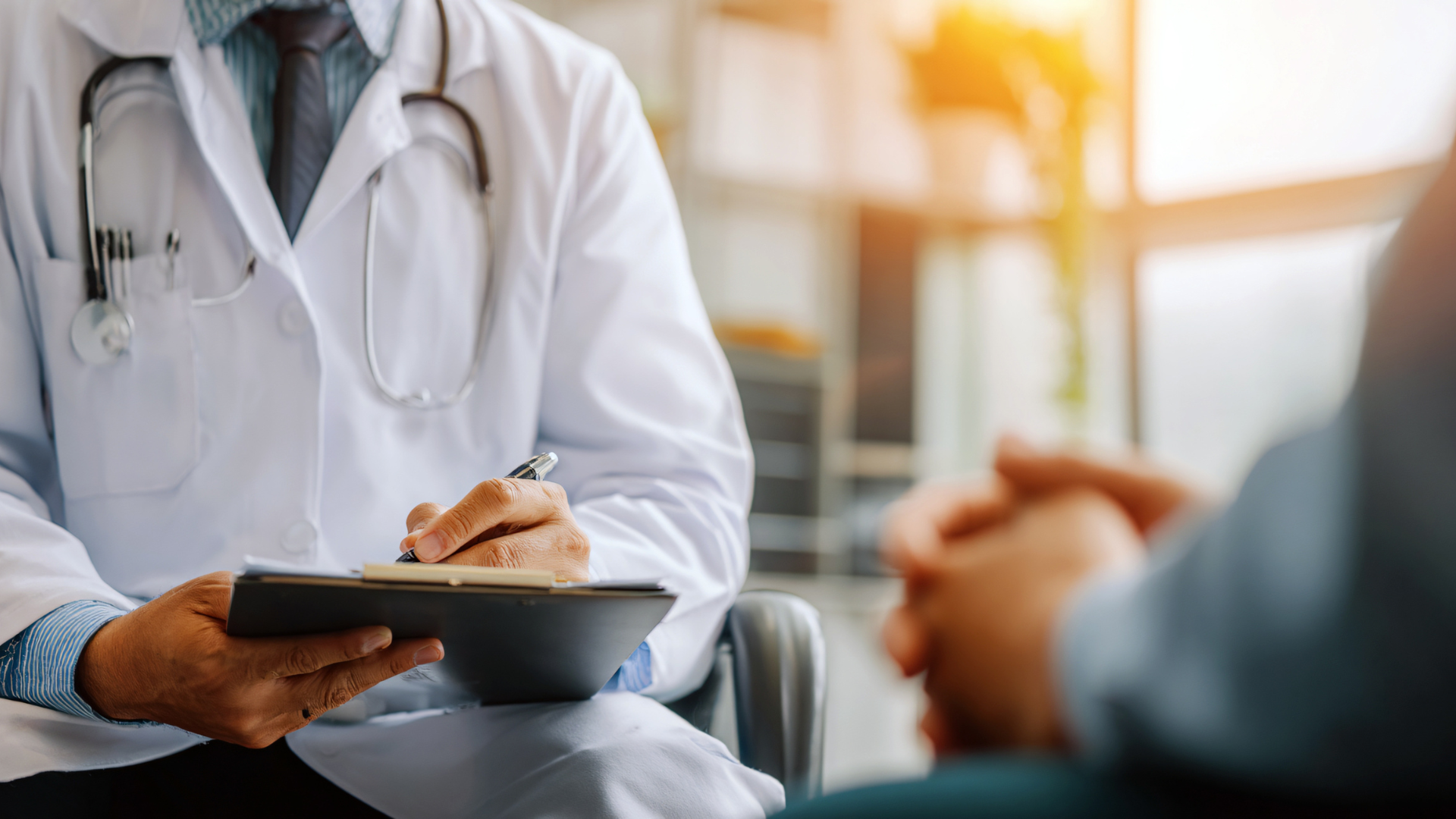 Doctor in white coat writing on clipboard, consulting with a patient in an office.