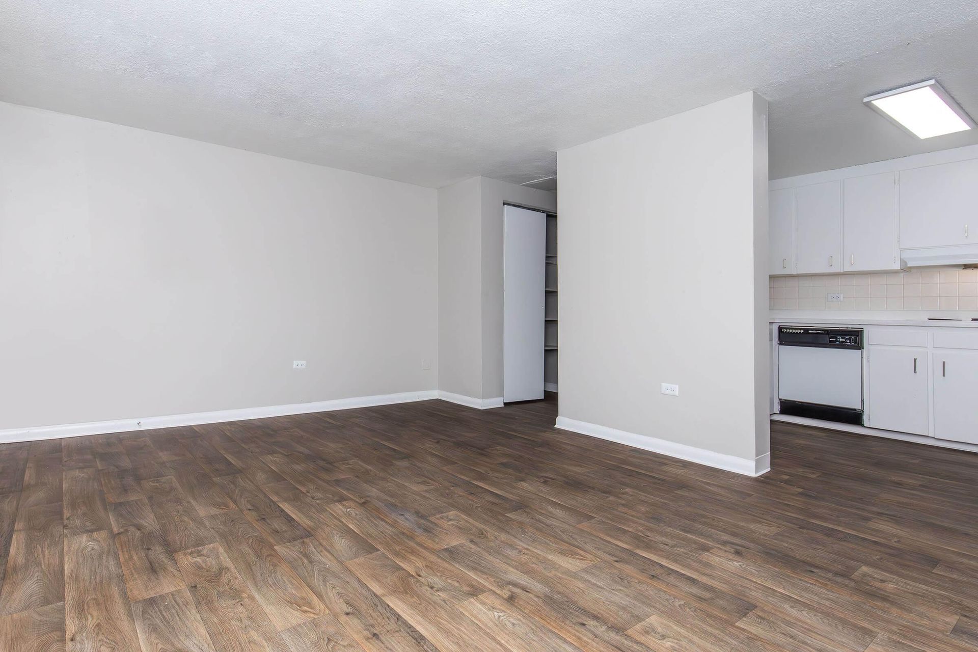 Empty apartment interior with wood flooring, light gray walls, and white kitchen cabinets.