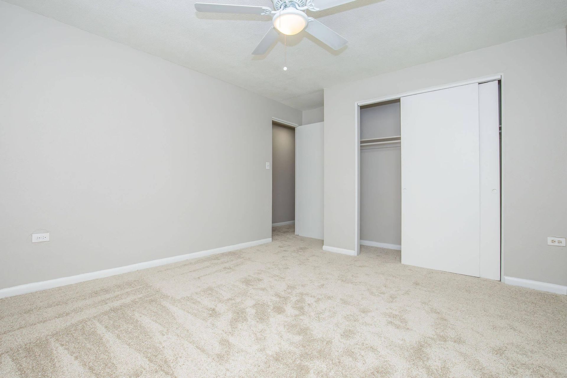 Empty bedroom with beige carpet, light gray walls, white closet doors, and ceiling fan.