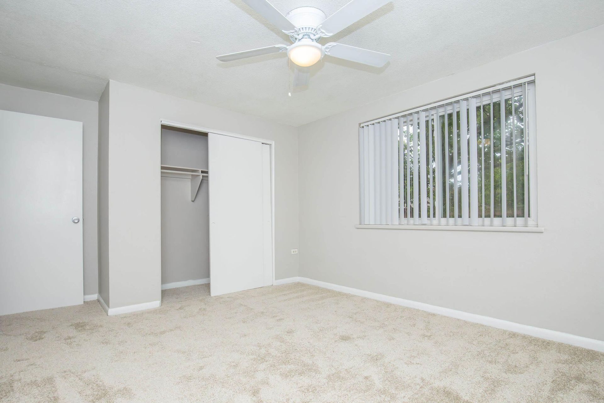 Empty bedroom with closet, window, ceiling fan; beige walls, carpet.