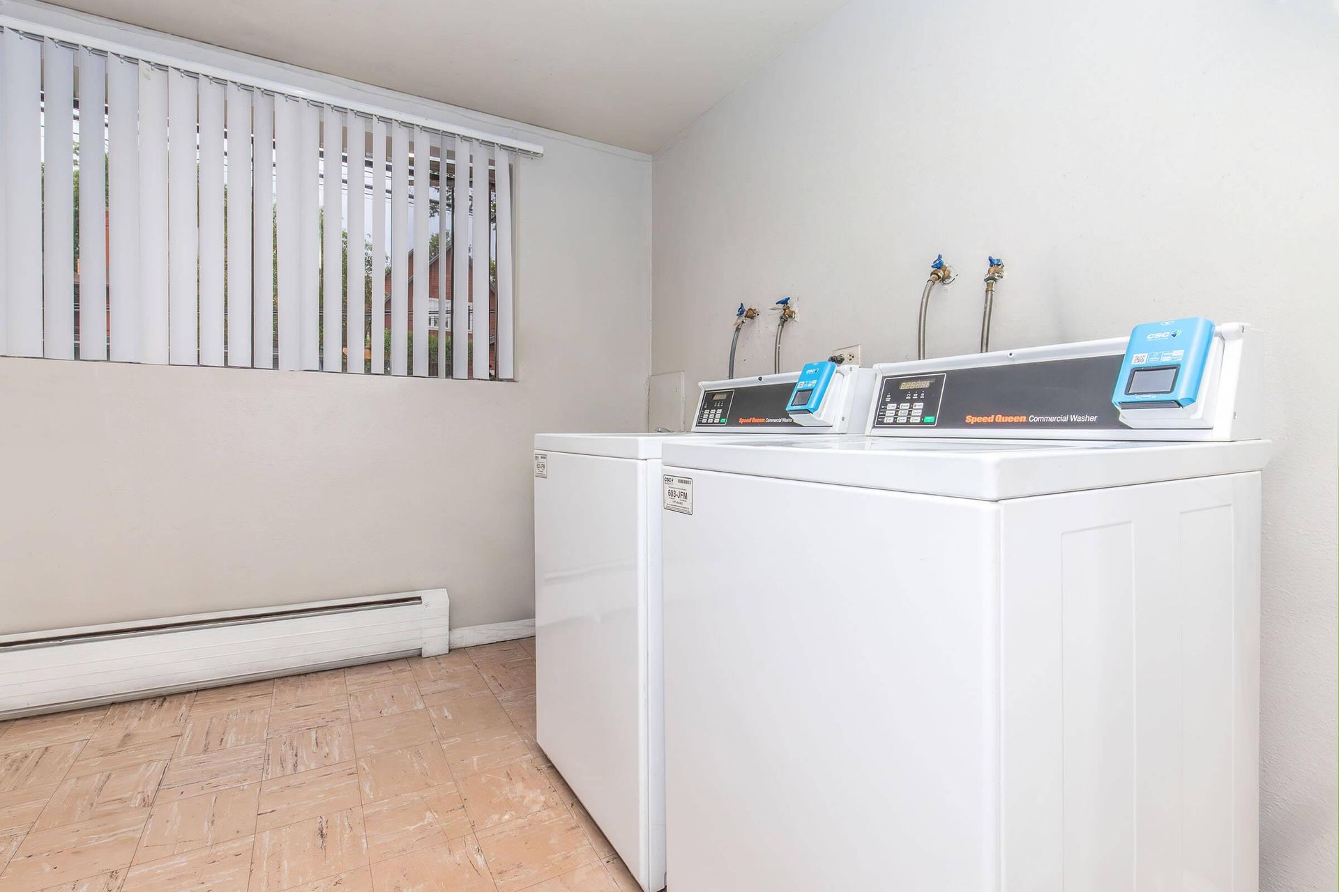 Laundry room with two white washing machines, window with blinds, and beige flooring.