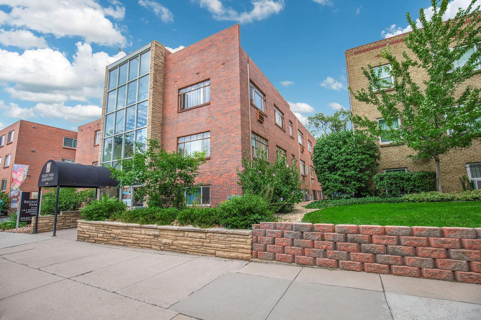 Brick apartment building with glass facade, green landscaping, and blue sky.