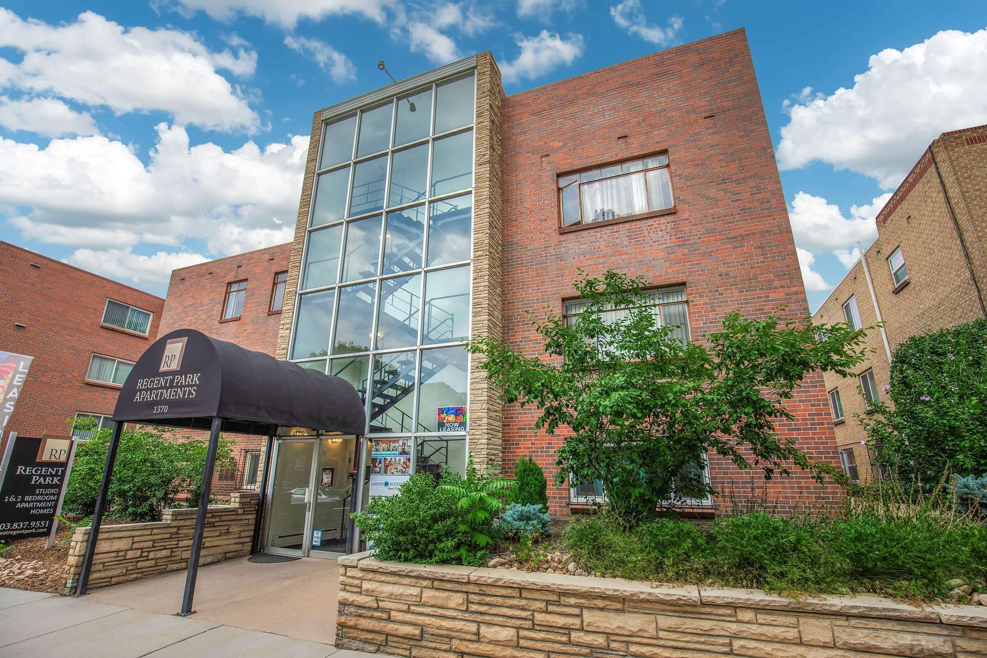 Brick building with glass entrance, arched awning, and greenery under a blue sky.