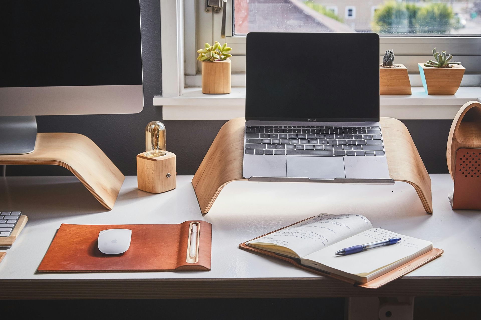 Laptop on a wooden stand, desk setup with monitor, mouse, notebook, and succulents by a window.