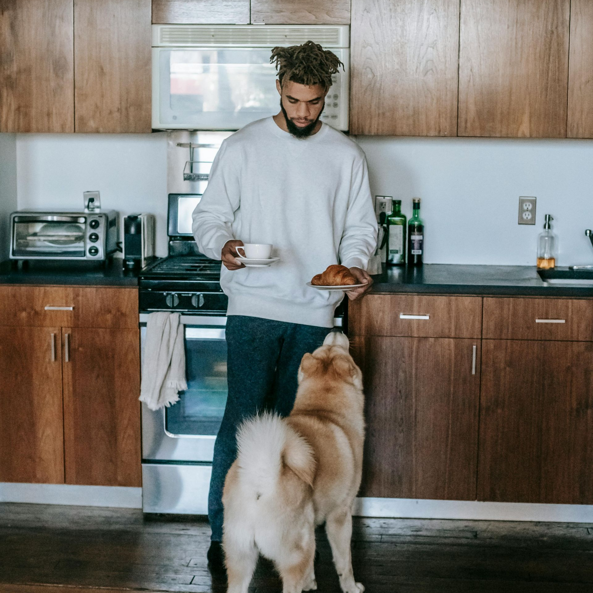 Man holding coffee and pastry in kitchen, dog looking up, waiting for food.